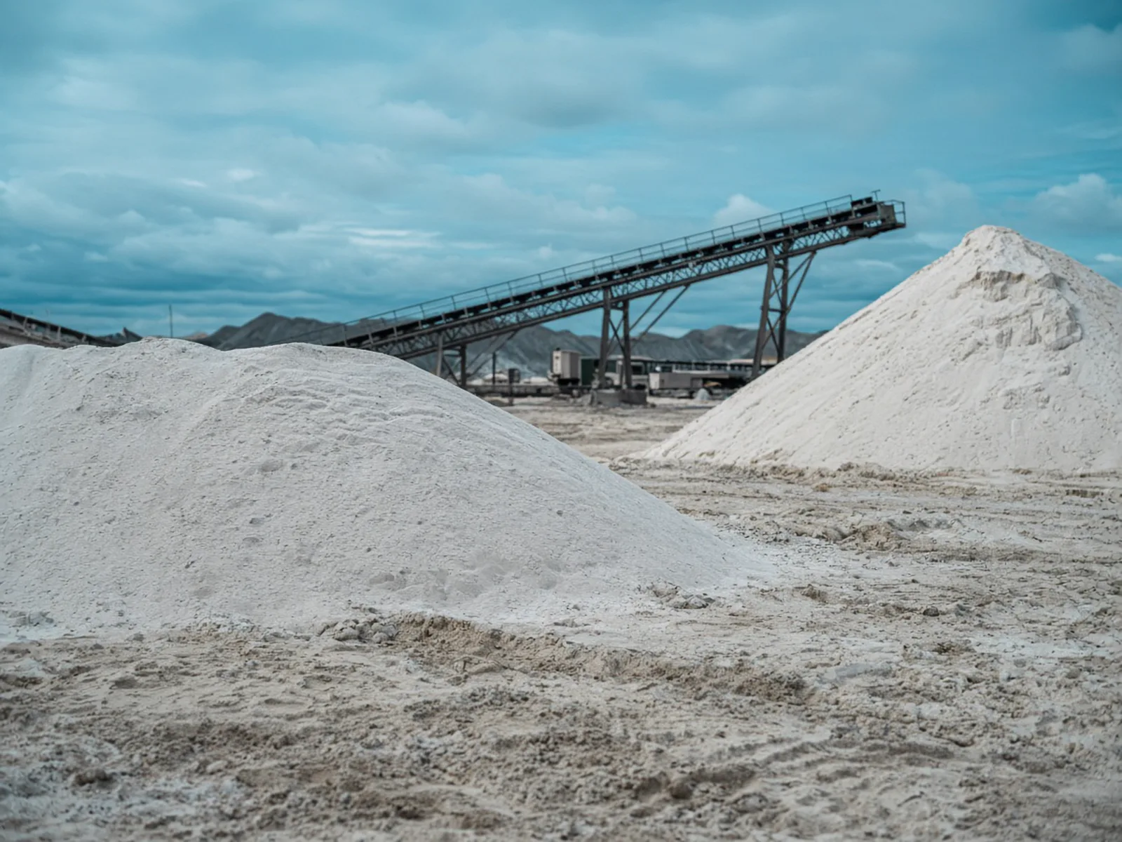 White mineral piles in a processing yard, with a conveyor behind them